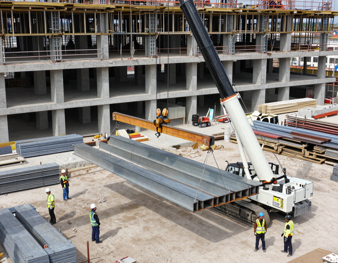 Obra en construcción con trabajadores guiando una grúa que levanta largas vigas de acero junto a la estructura de un edificio