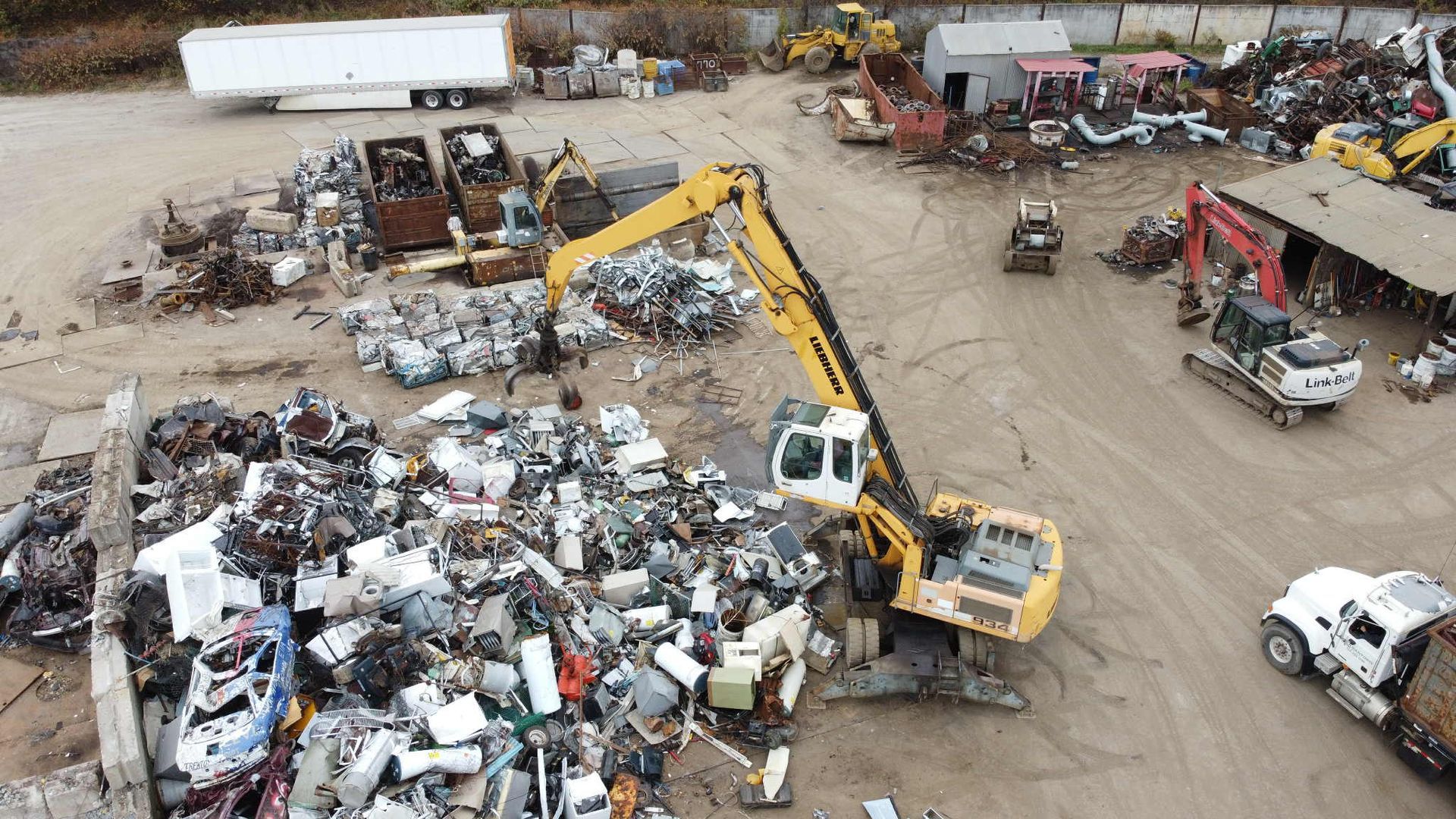 Large excavator sorting scrap metal at a junkyard. Other heavy machinery and trucks are also present.