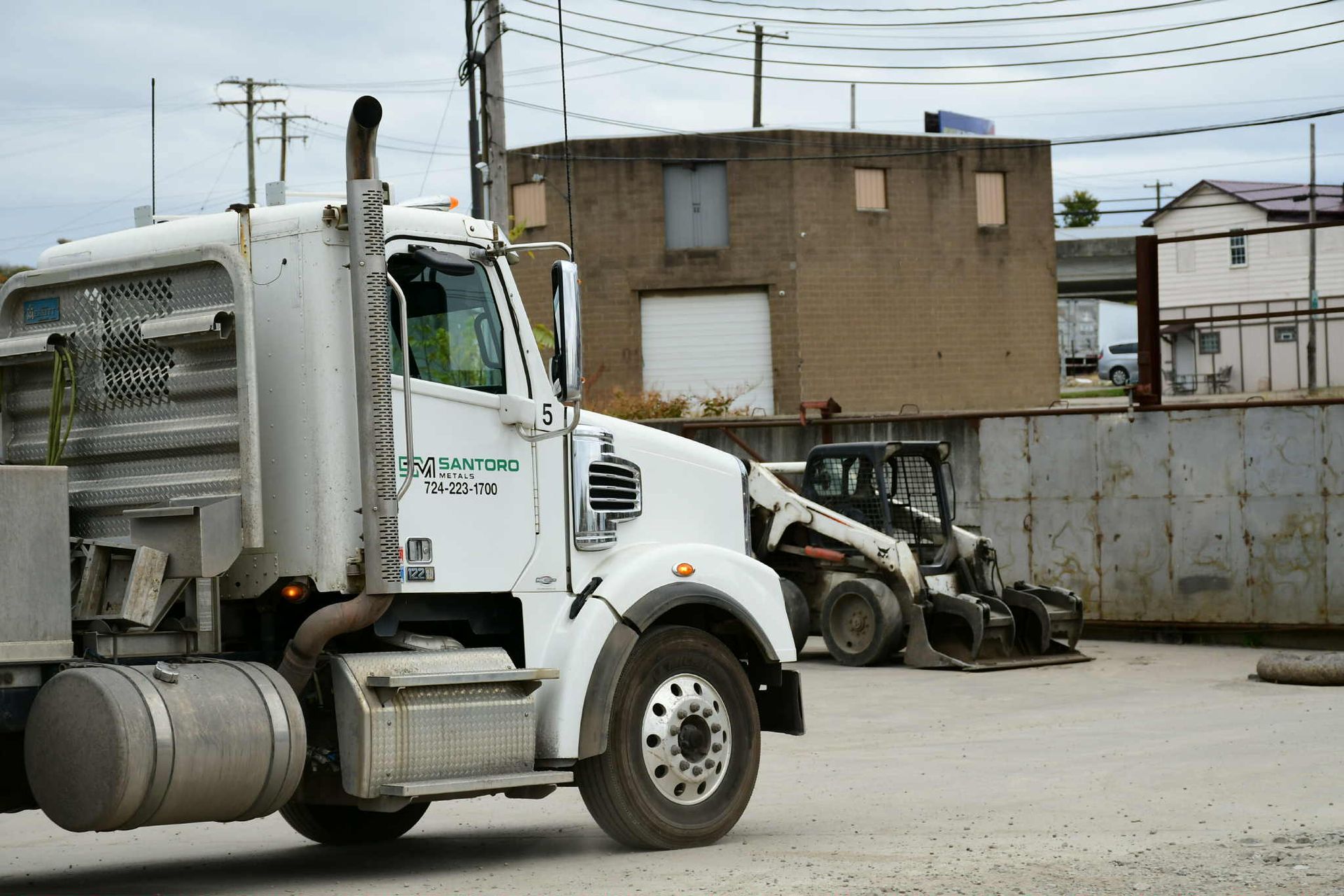 White semi-truck and Bobcat loader at a construction site near a tan building.