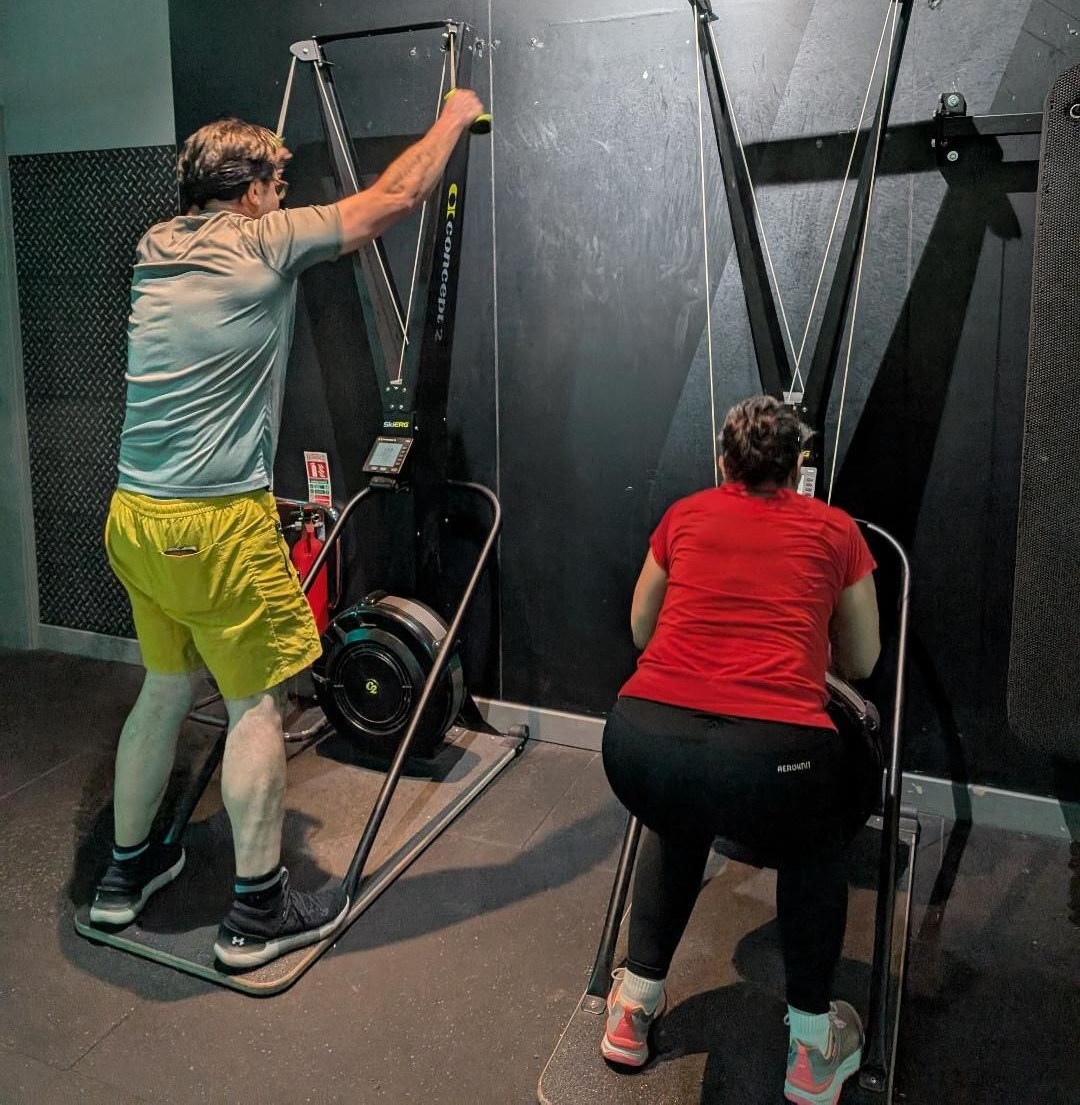 Two people using ski machines in a gym. Man in yellow shorts and woman in red shirt work out with equipment.