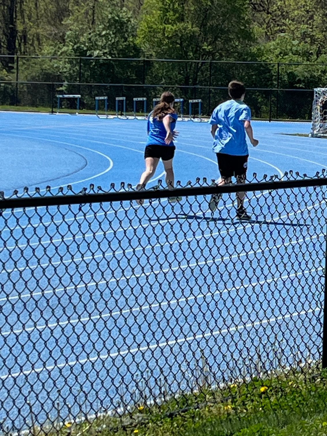 Two people are running on a track behind a chain link fence