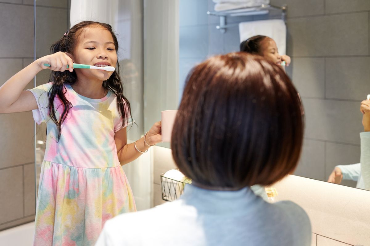Child brushing teeth in a bathroom, seen in the mirror while an adult watches nearby.