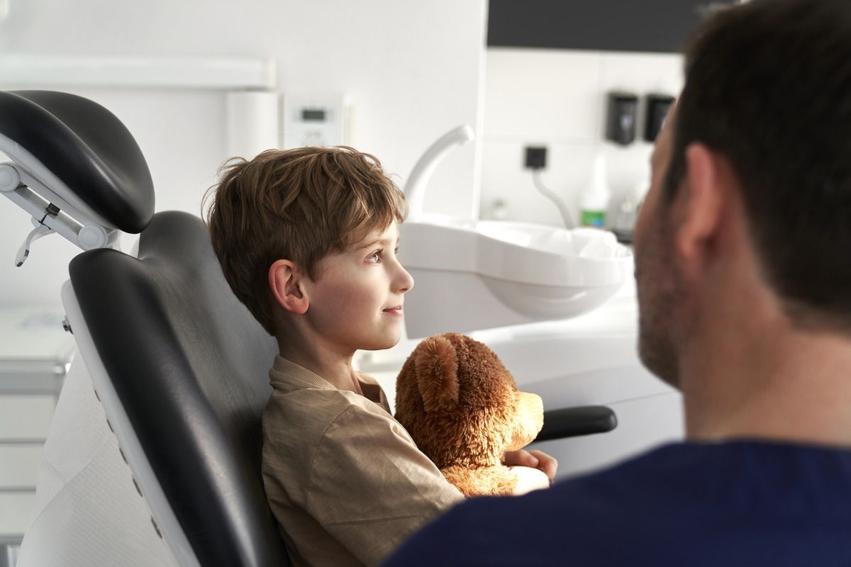 A young boy is sitting in a dental chair holding a teddy bear.