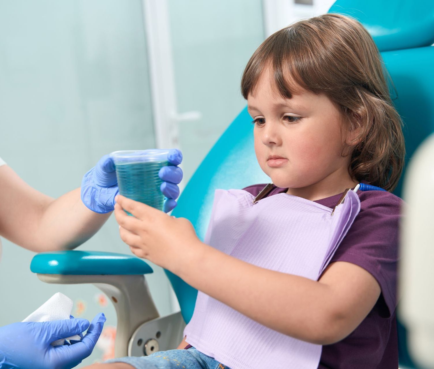 A little girl is sitting in a dental chair holding a cup of liquid.