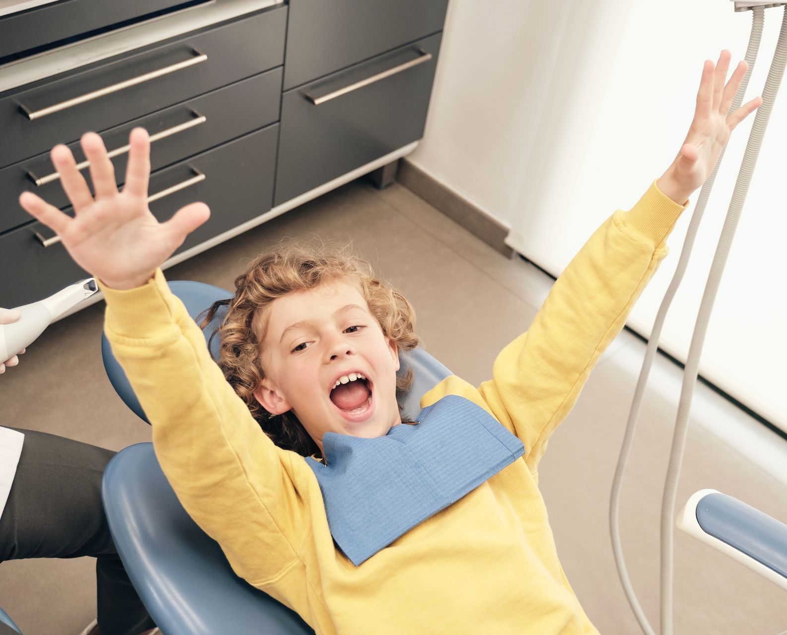 A young boy is sitting in a dental chair with his arms in the air.