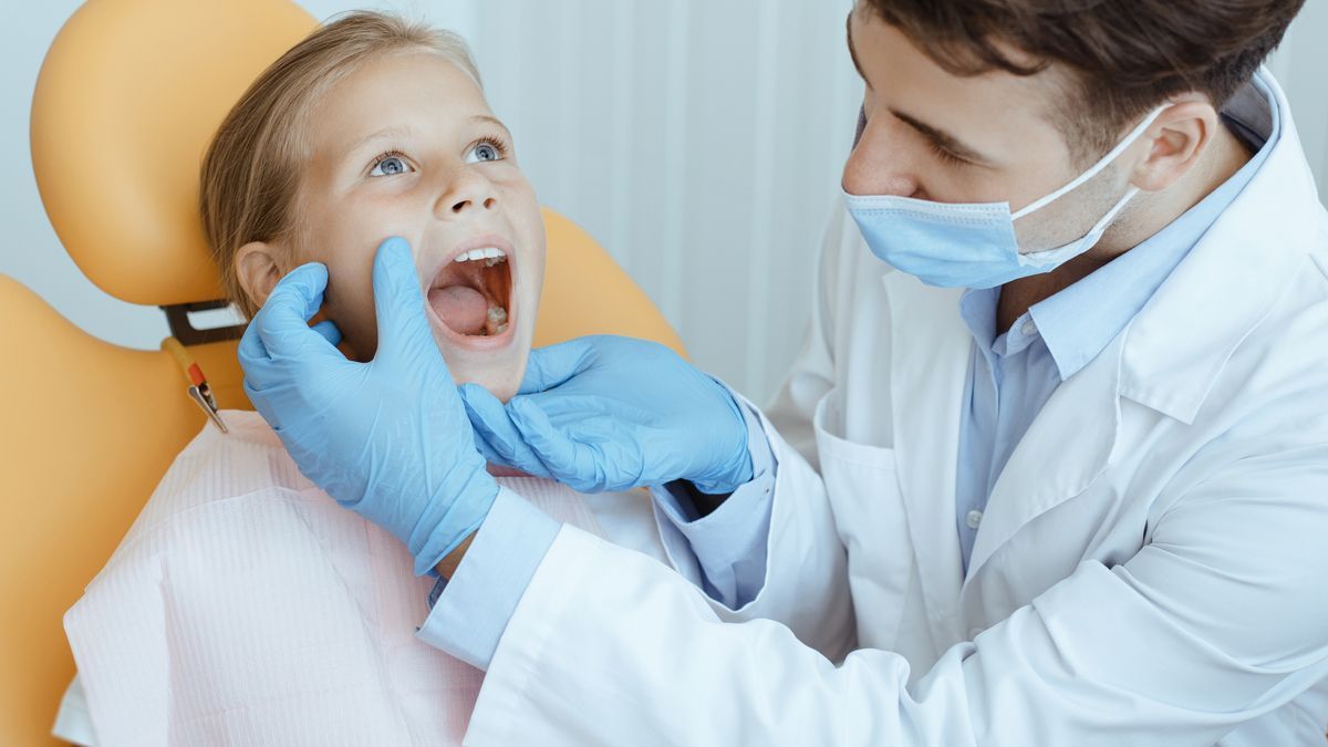 Dentist examining a child’s open mouth in a dental chair, wearing gloves and a mask.