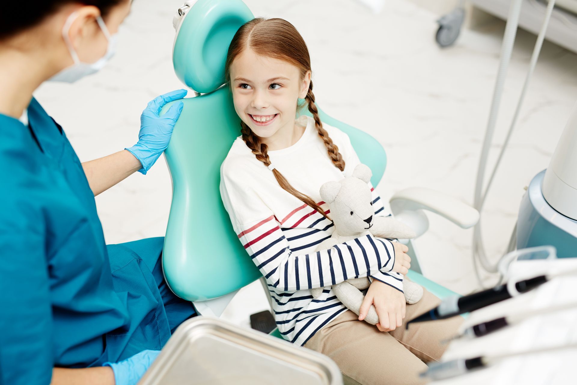 A little girl is sitting in a dental chair holding a teddy bear.