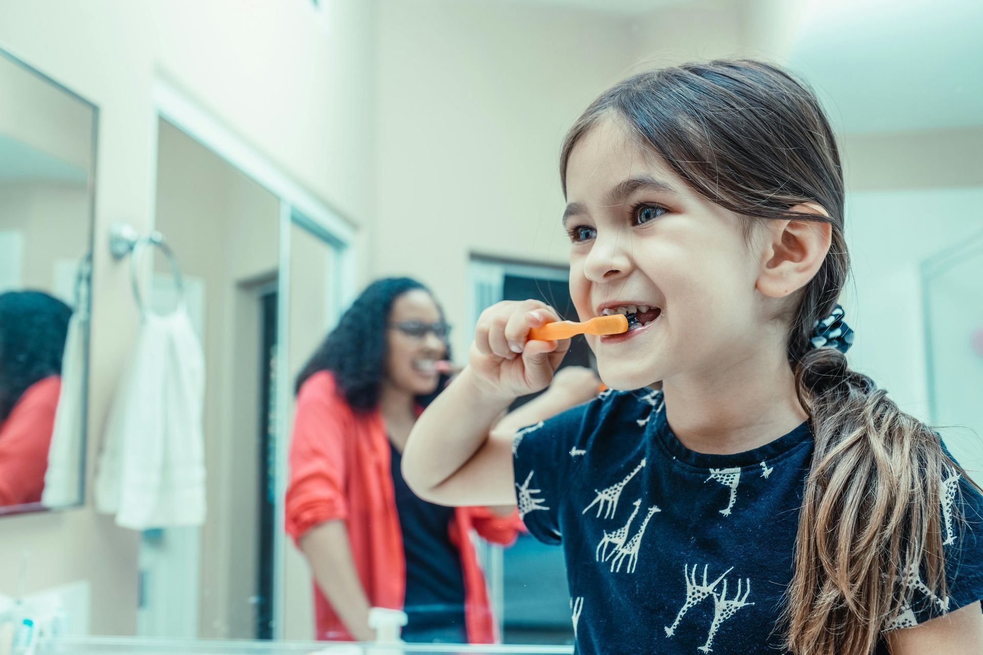 Child holding toothbrush, brushing teeth in mirror with another woman showing how to brush teeth
