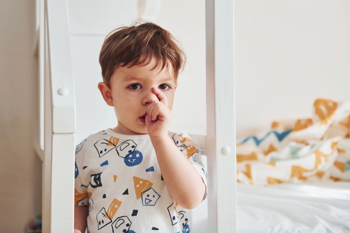 Young child in a patterned shirt standing by a bed, finger in mouth, in a bright bedroom
