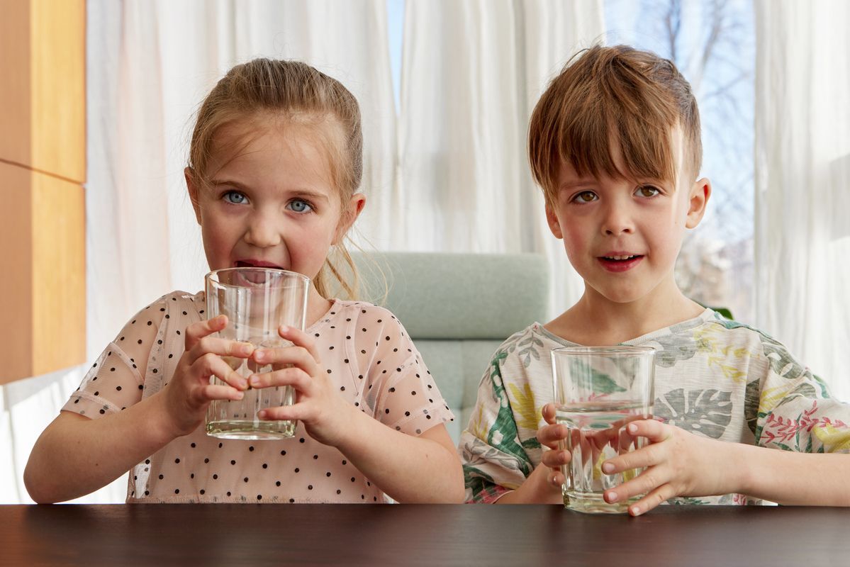 Two children at a table holding glasses of water, with a bright window in the background