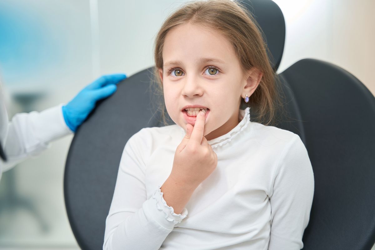 Child sitting in a dentist’s chair, touching a tooth while a gloved hand rests nearby