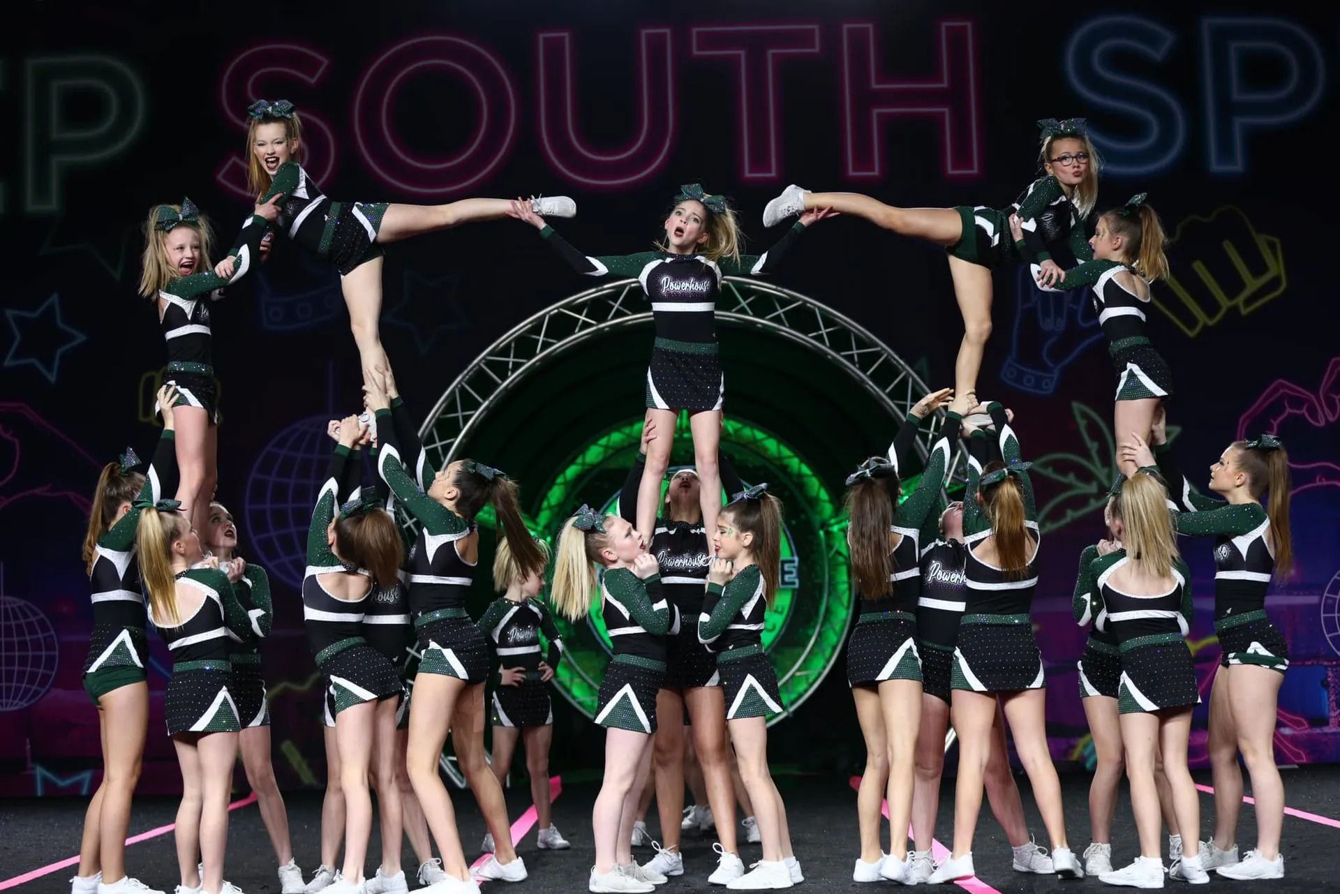 Cheer squad performing a stunt on stage, with athletes lifted in green and black uniforms under purple lighting