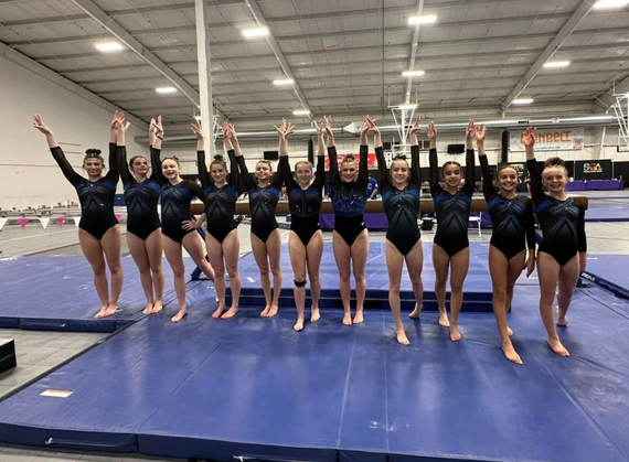 Gymnastics team posing with arms raised on a blue mat inside an indoor training gym.