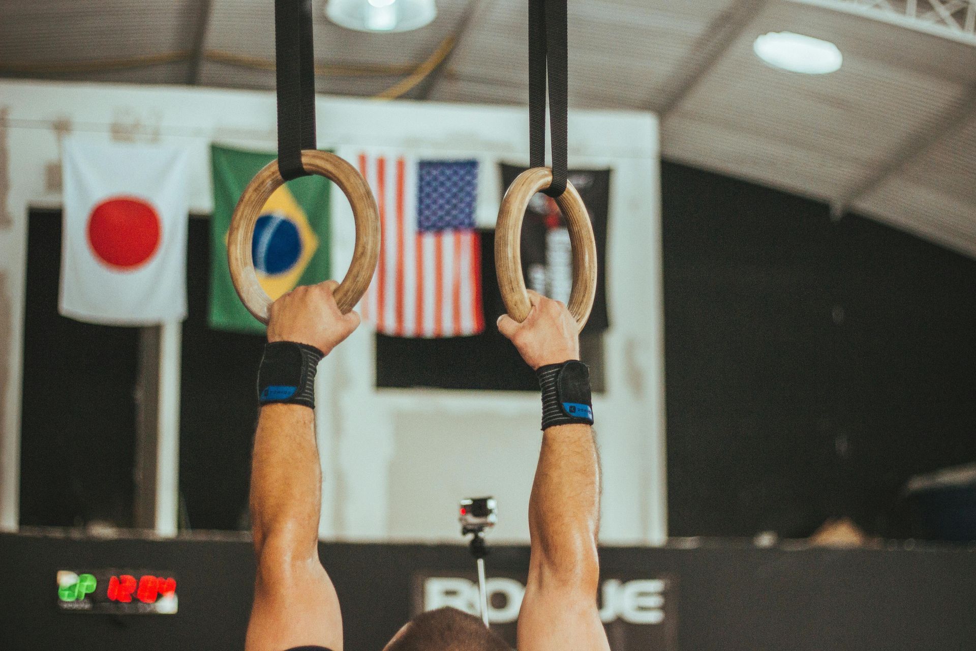 Hands gripping gymnastic rings in a competition arena with national flags in the background