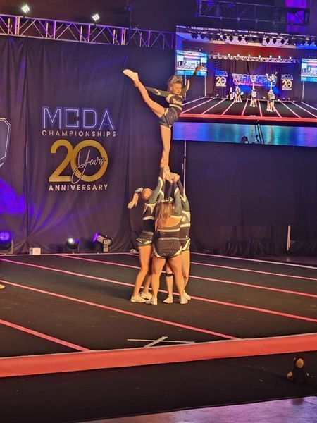 Cheerleading stunt on stage, with teammates holding a flyer aloft under purple lights at MCDA Championships