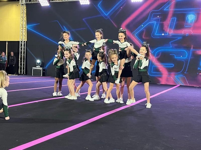 Cheerleading team posing on a stage with neon blue and pink lights, arms raised in a group formation