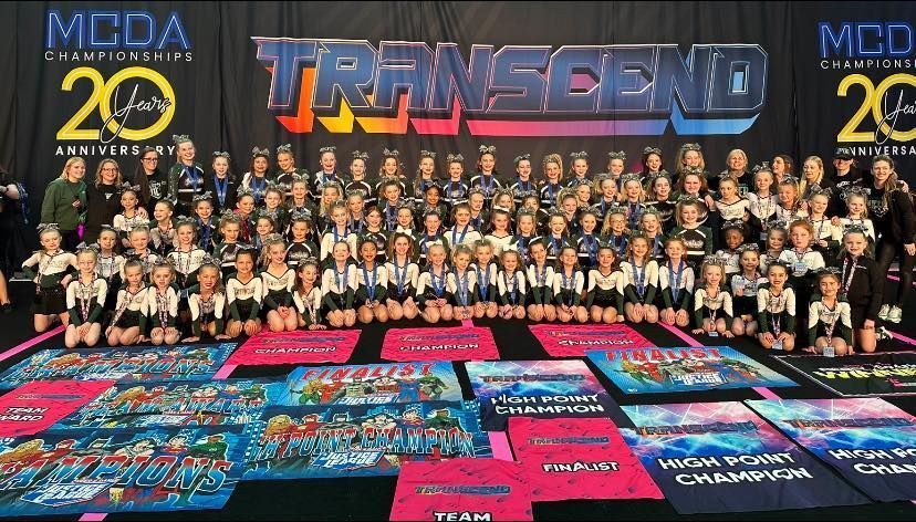 Large group photo of performers and crew on stage with a “TRANSCEND” backdrop and colorful awards banners