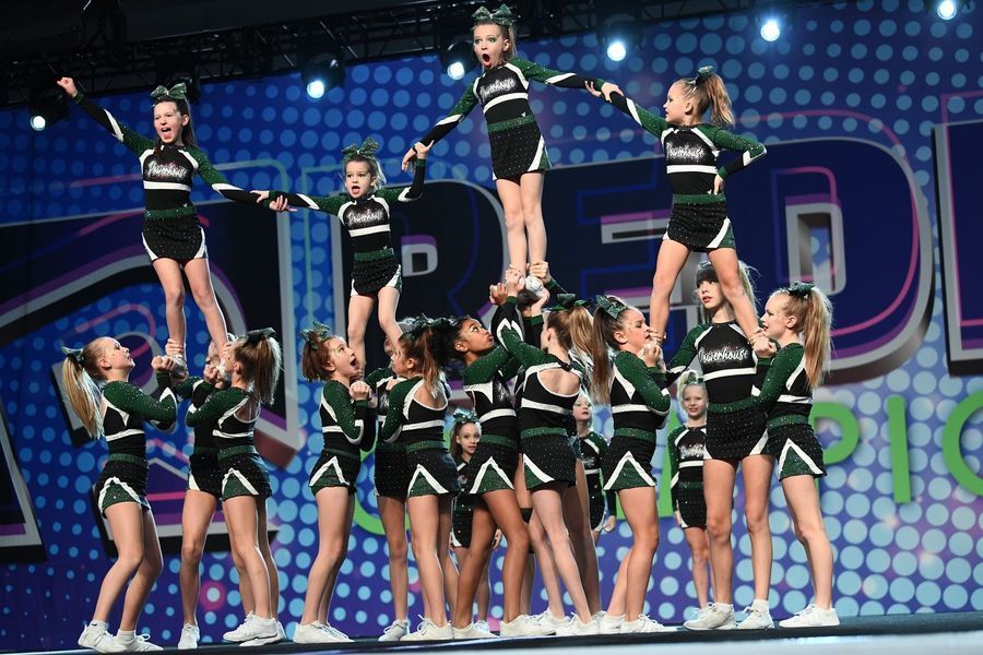 Cheerleaders in black and green uniforms perform stunts on a stage with blue lights and a dotted backdrop