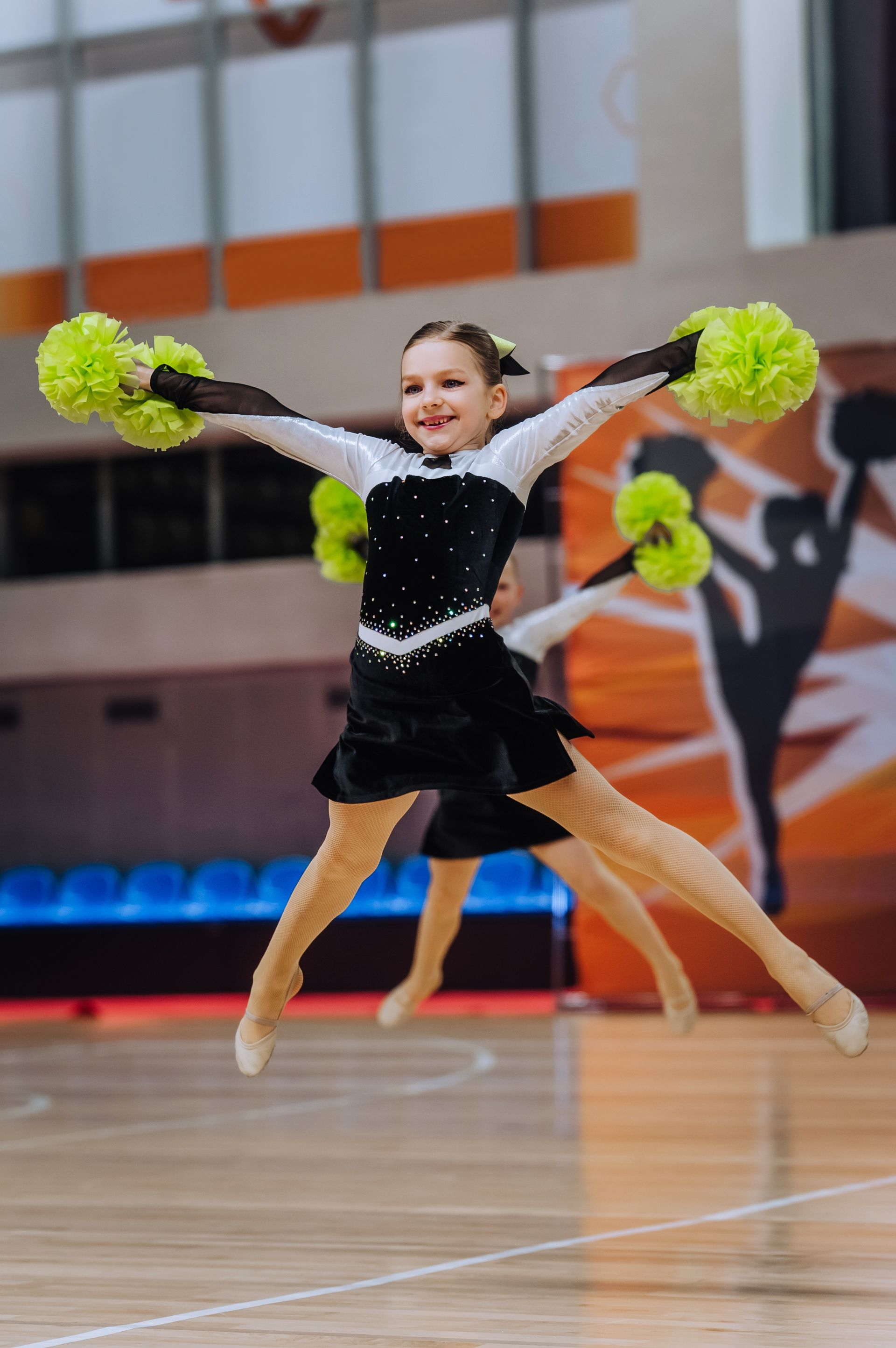 Cheerleader mid-jump on indoor court, holding neon green pom-poms and wearing a black costume.