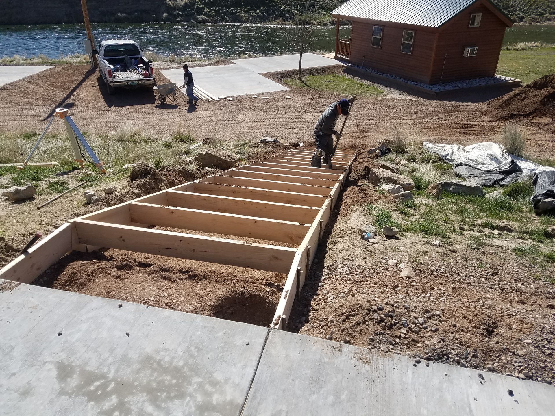 Workers building wooden stairs on a sloped yard beside a lake and cabin, with a truck nearby