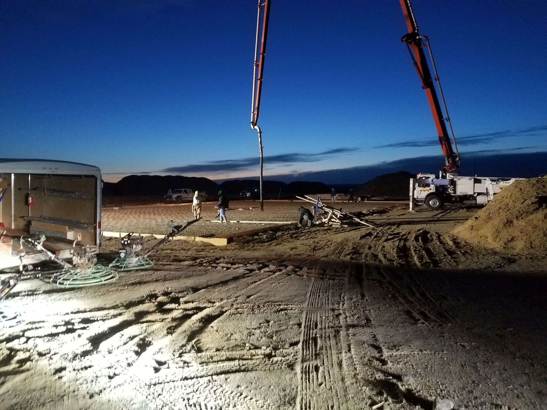 Construction site at dusk with cranes, dirt piles, and gravel road under a blue sky