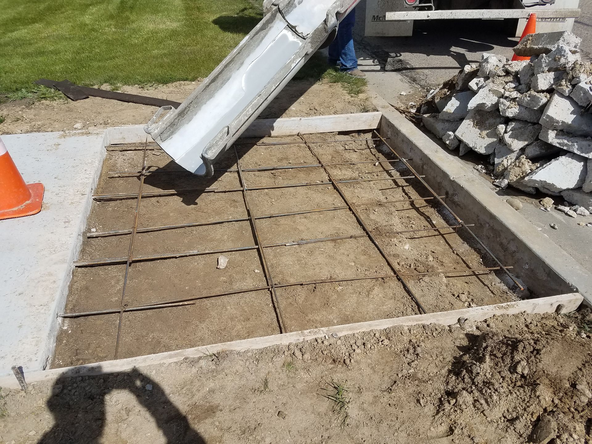Freshly leveled dirt pad with a concrete border, pipe, rubble pile, and orange cone near a curb.