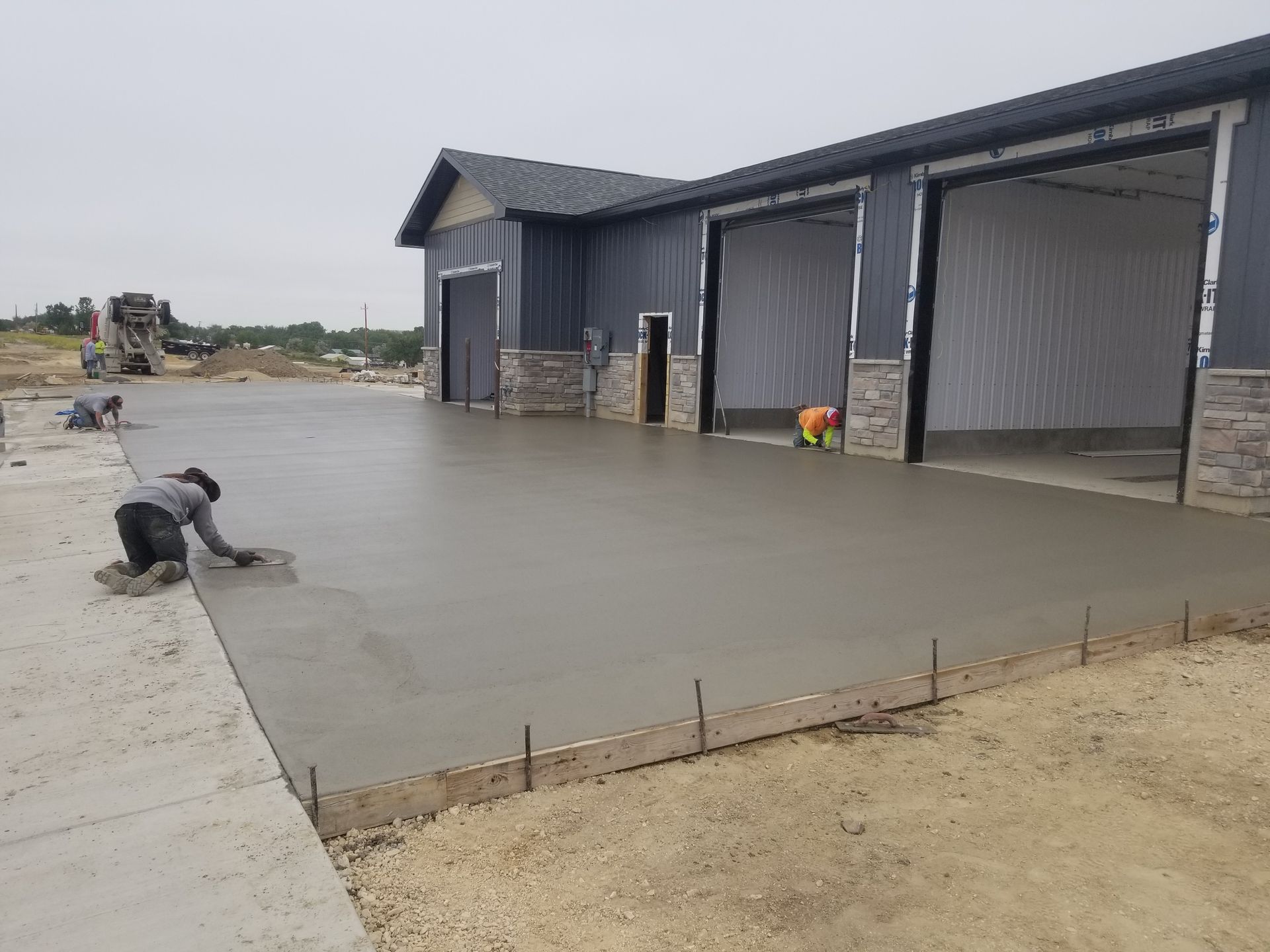 Worker smoothing fresh concrete beside a large metal building under construction