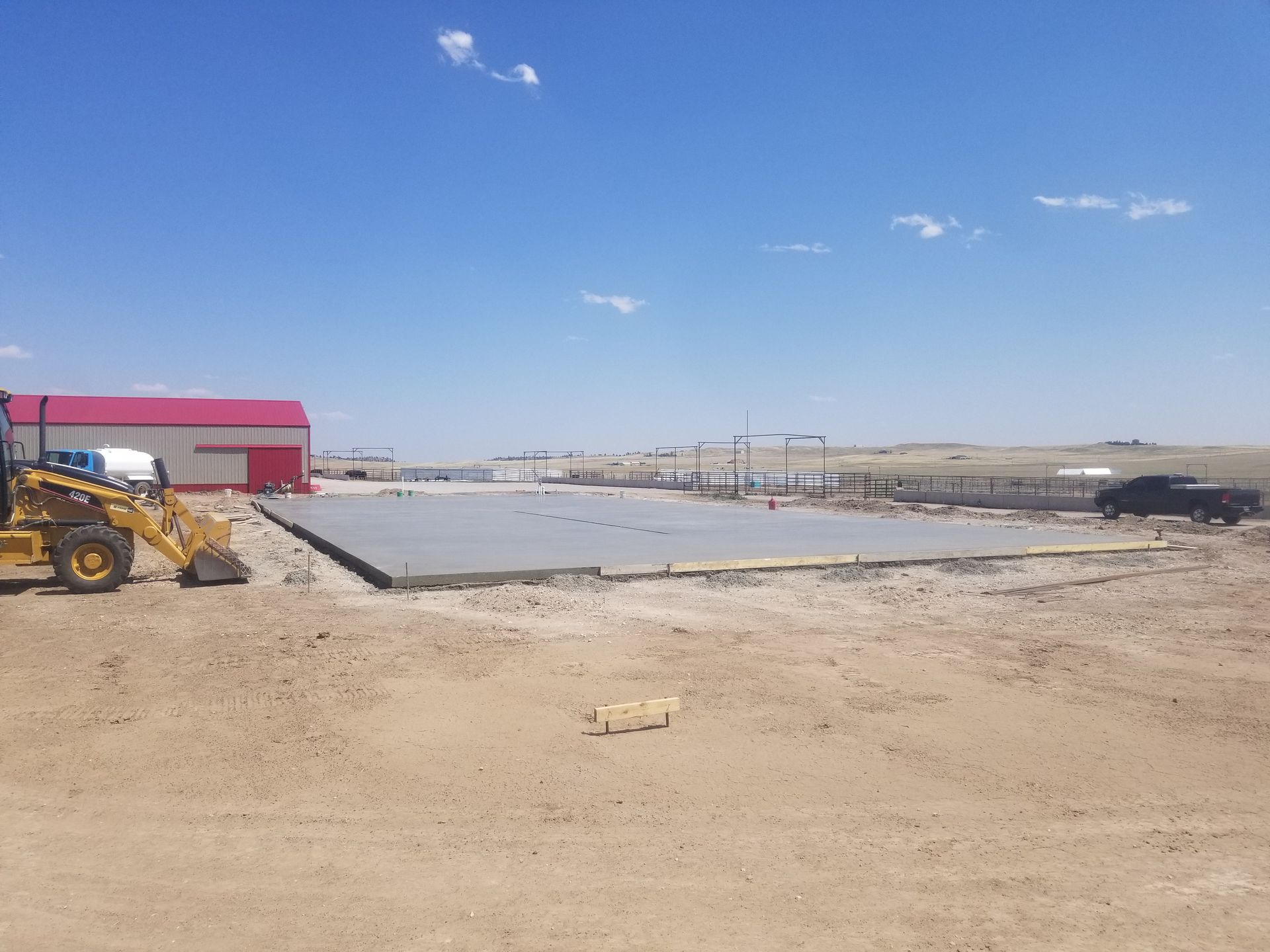 Construction site with a concrete slab, yellow loader, and red-roofed building under a clear blue sky