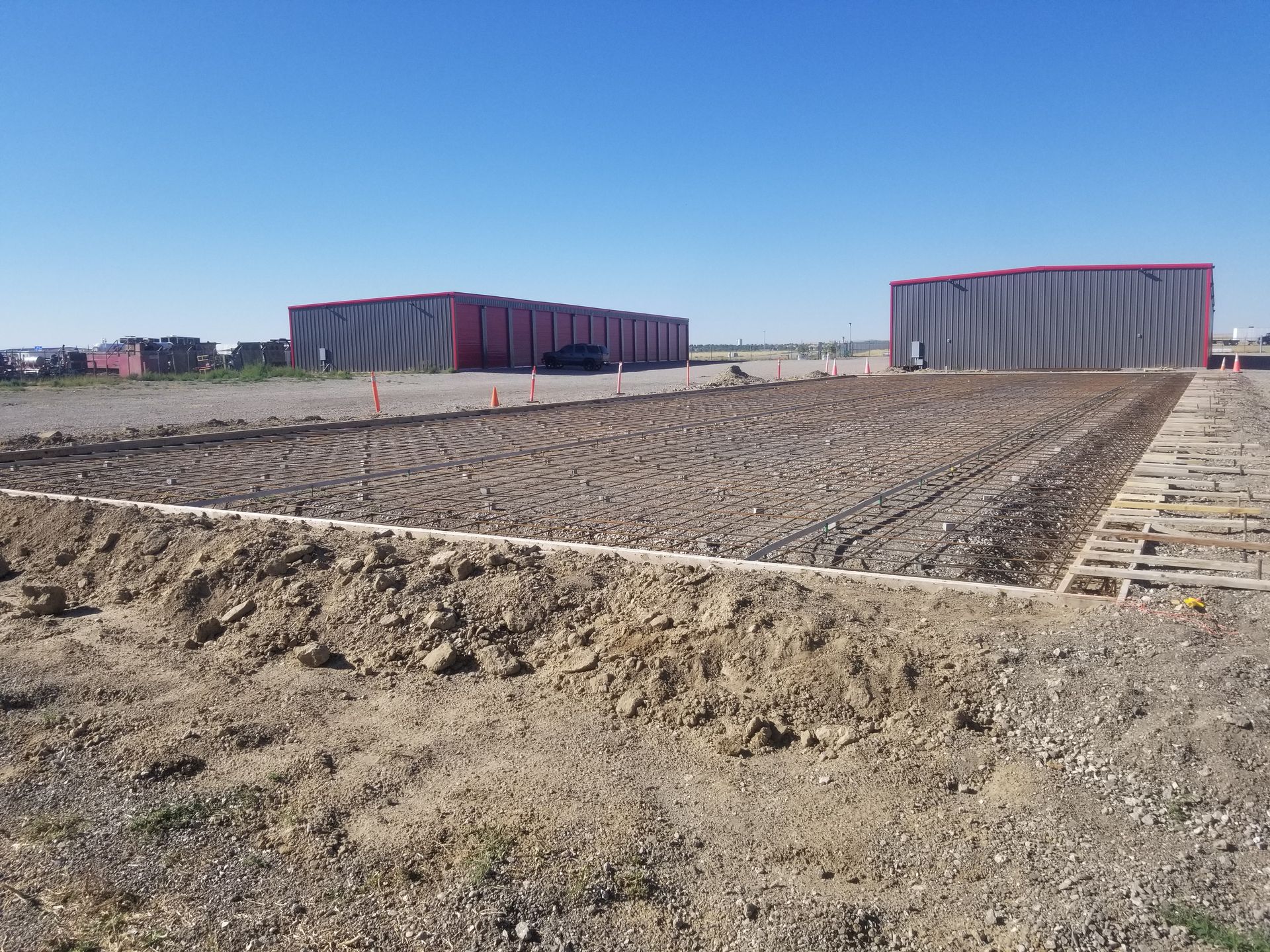 Bare dirt construction site with two large gray and pink industrial buildings under a clear blue sky