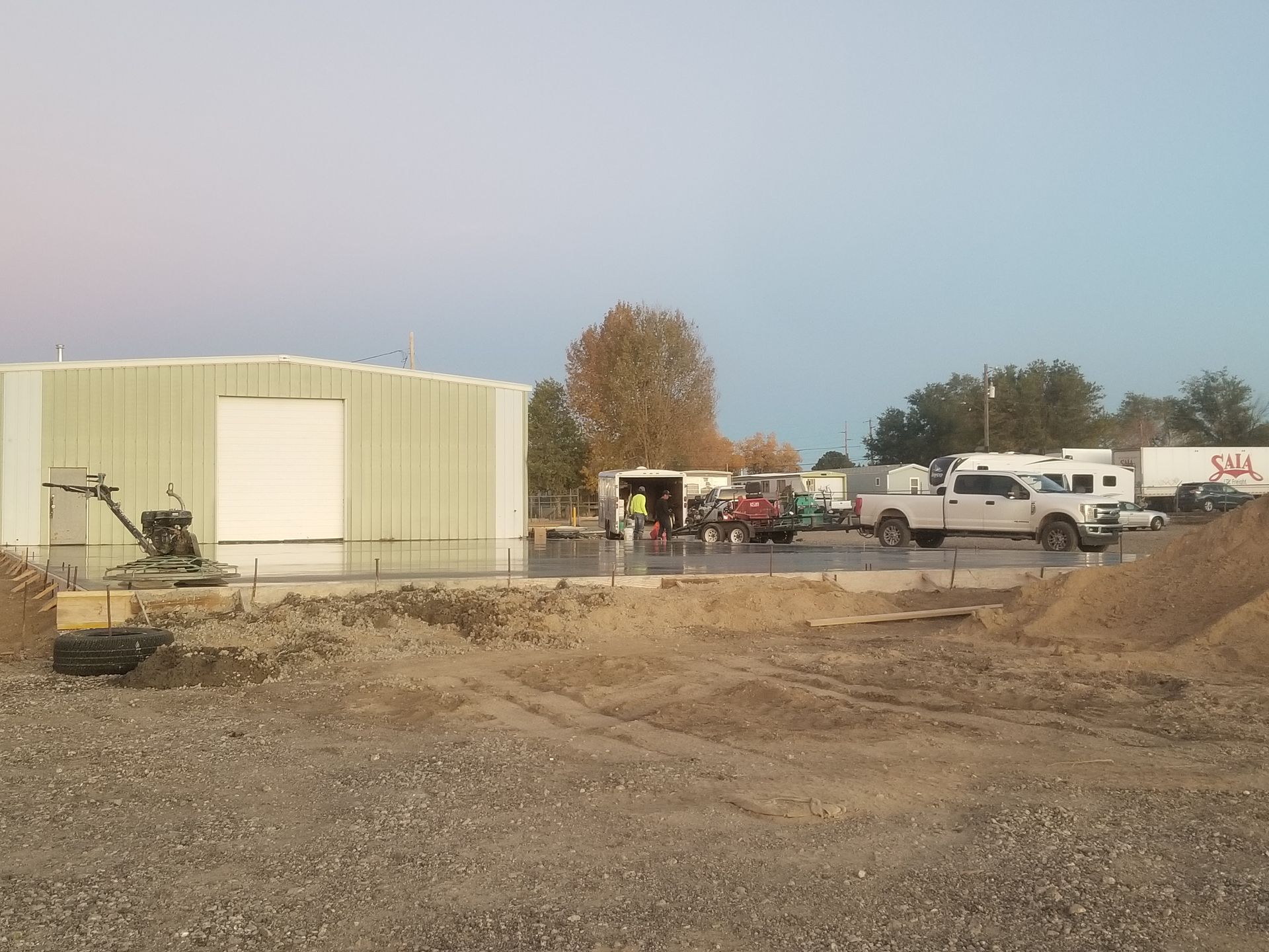 Construction site with a large metal building, parked trucks, and equipment on a dirt lot under a cloudy sky