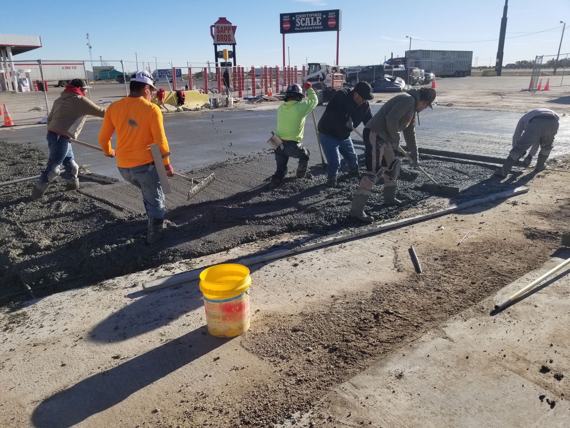 Road crew spreading asphalt on a damaged street beside a highway, with a yellow bucket in the foreground