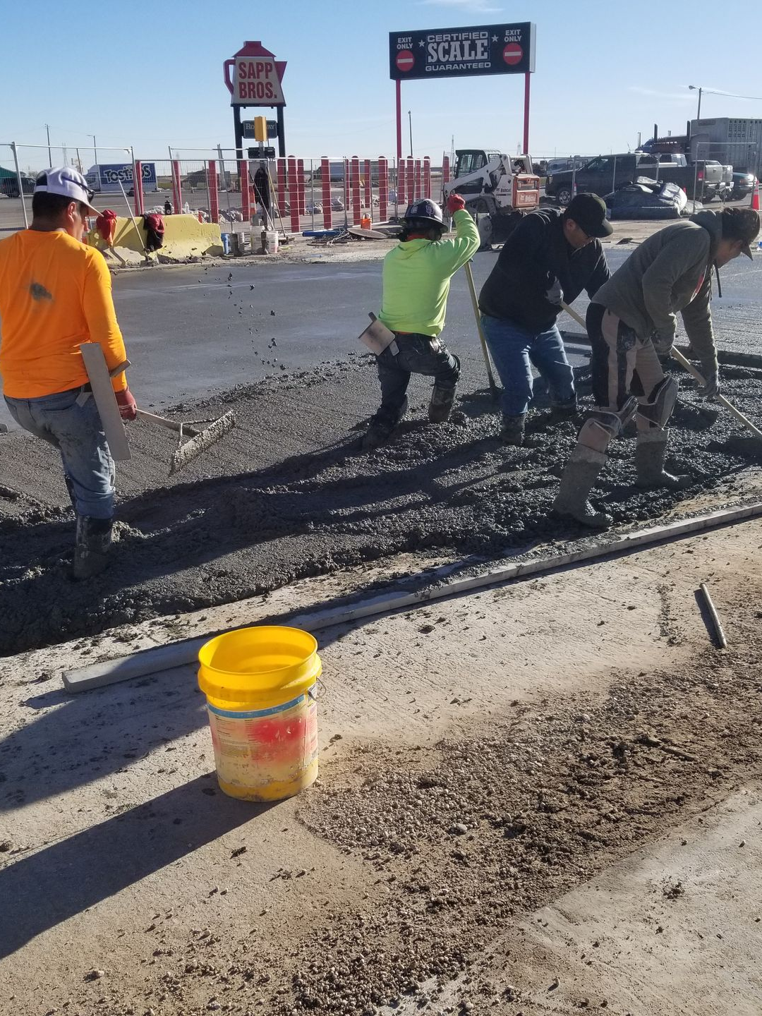 Workers patching a cracked road beside a yellow bucket at a roadside construction site.