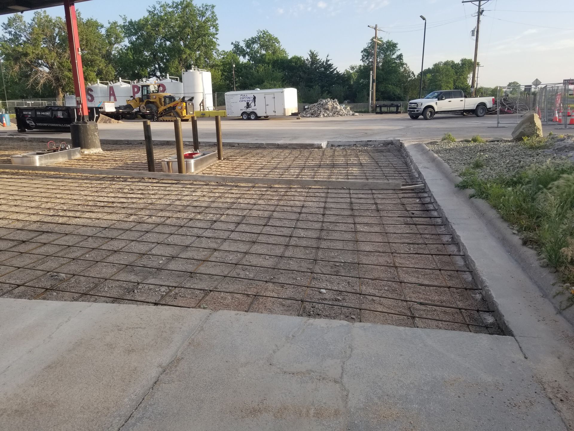 Empty parking lot with paved and gravel surfaces, trucks in the distance, and trees along the edge