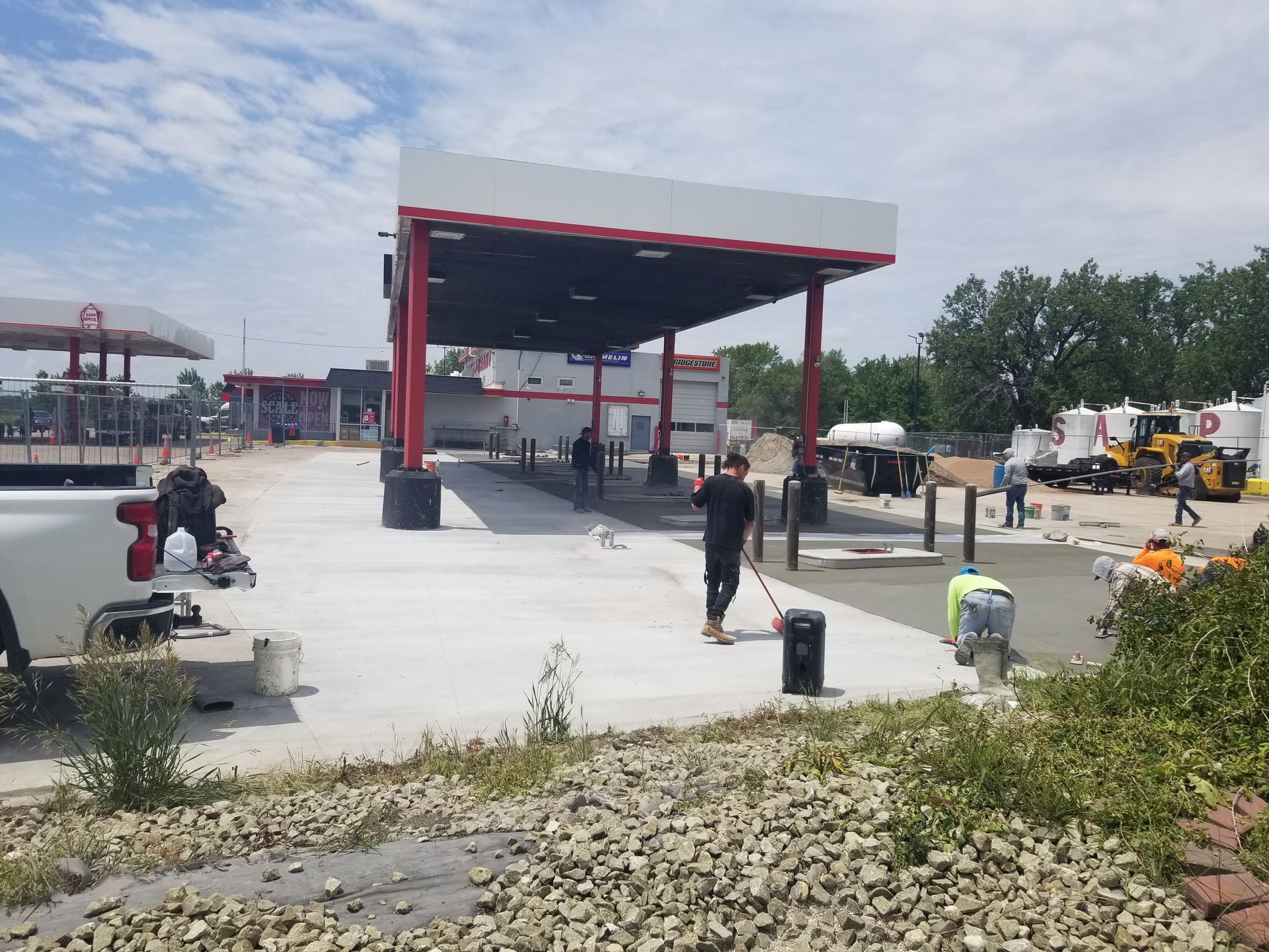 Workers under a red-roofed shelter at an outdoor construction site with equipment and concrete pavement.