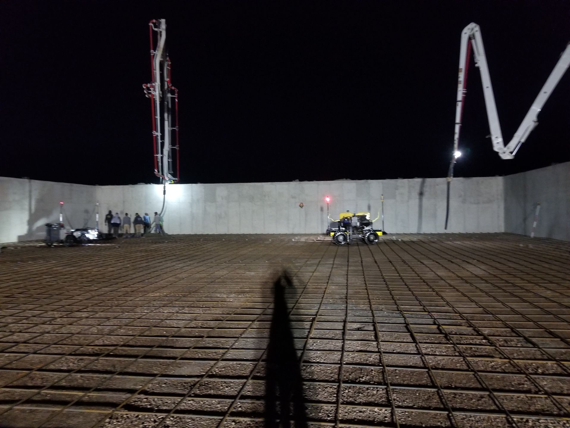 Construction site at night with concrete wall, rebar grid, and workers using machinery under cranes.