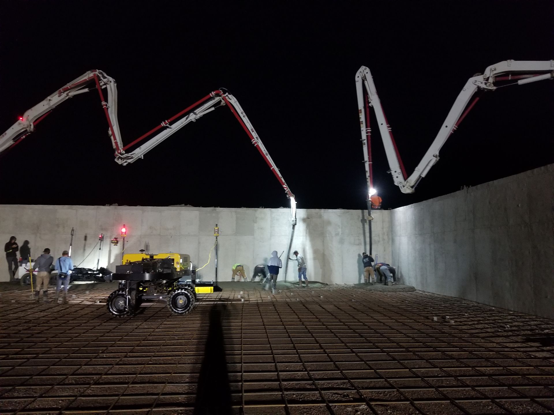 Construction workers pouring concrete in a large foundation at night with two boom pumps overhead