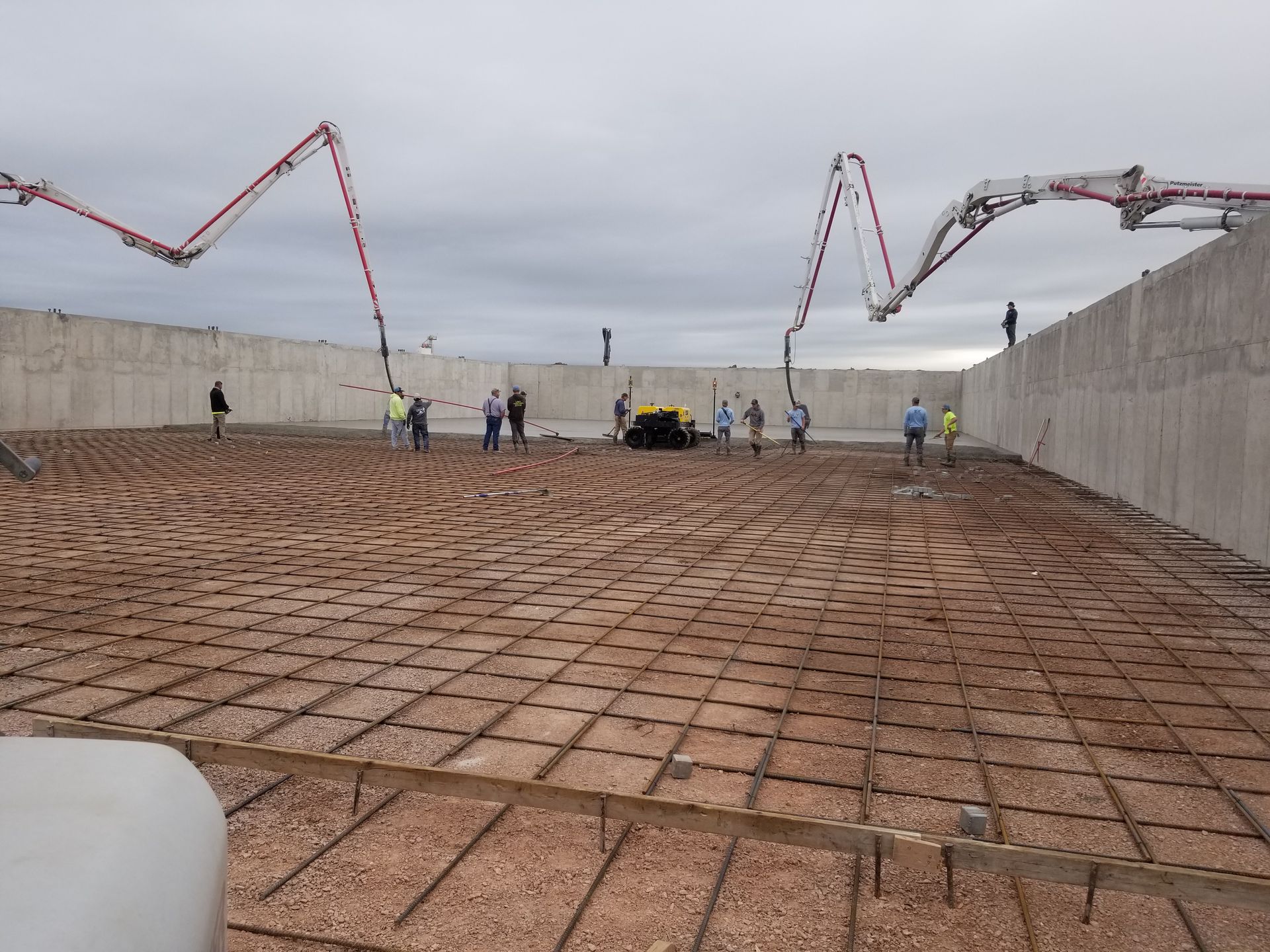 Construction workers on a sloped concrete surface with rebar, cranes, and retaining walls by the water