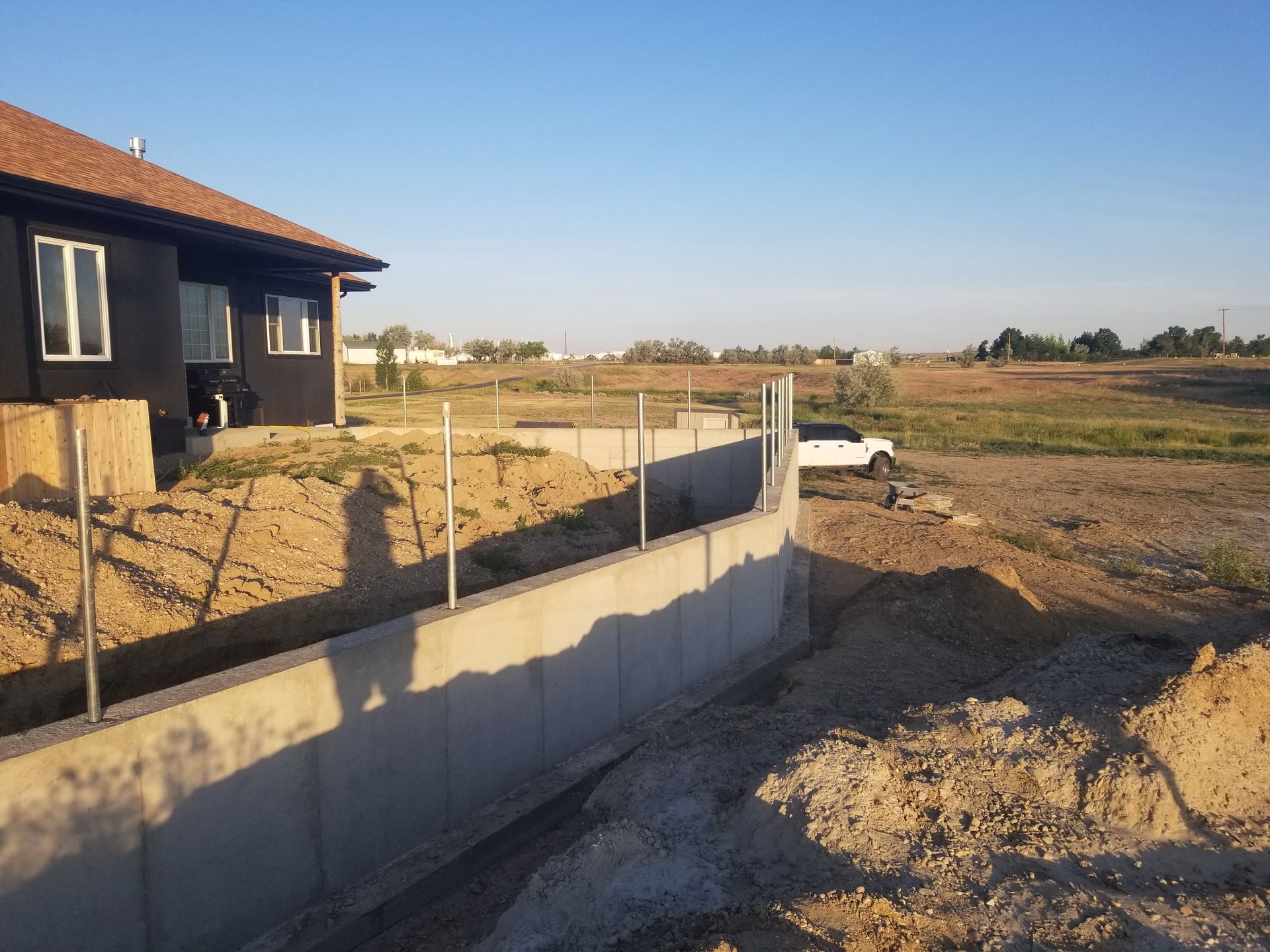 Partially built concrete wall beside a house on a dirt lot under a clear sky