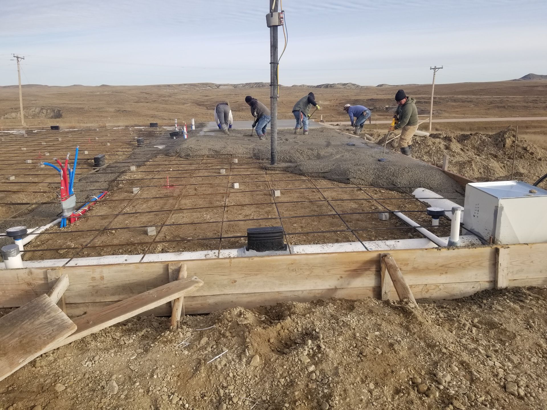 Workers pouring concrete for a foundation on a rural construction site with exposed rebar and forms.