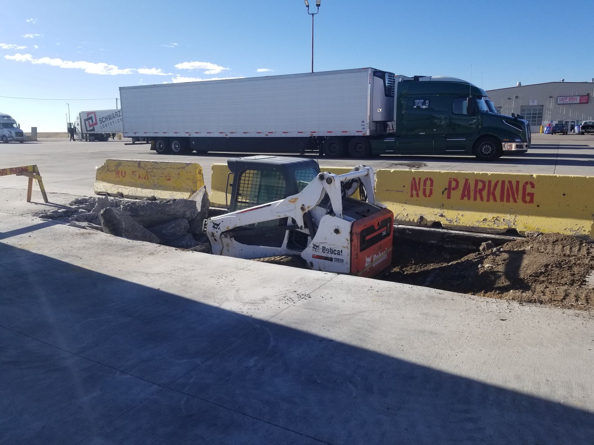 Excavator trapped in a ditch beside a yellow “NO PARKING” barrier and parked semi truck on a construction site
