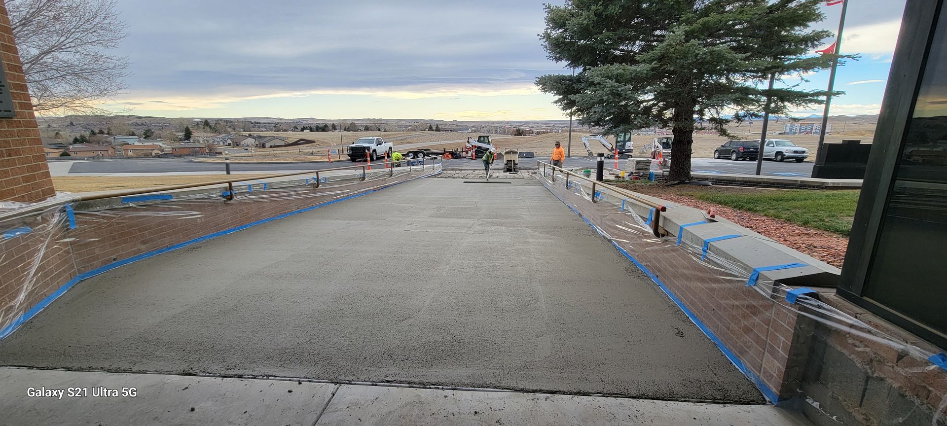 Concrete walkway to a beach with low walls, a tree, and distant ocean under a cloudy sky