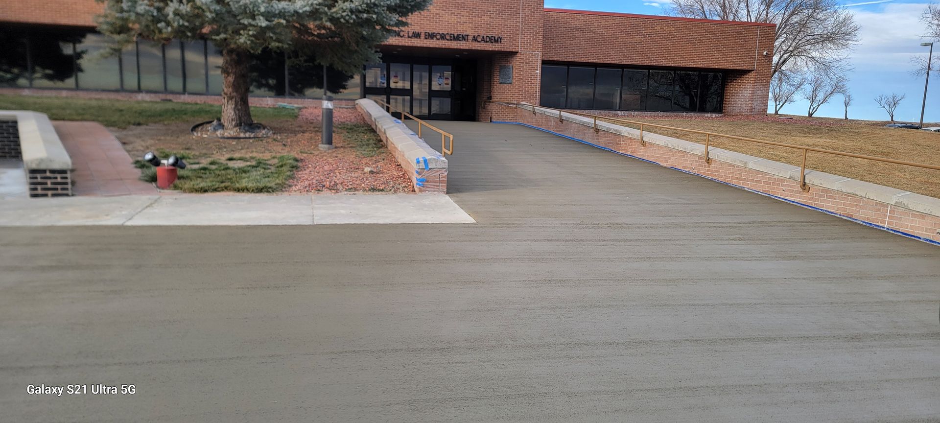 Large paved driveway leading to a brick building entrance with flower beds and a ramp.