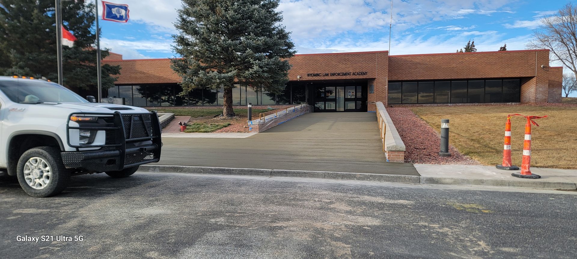 Driveway entrance to a low brick building, with a parked truck, trees, and orange traffic cones on the right.
