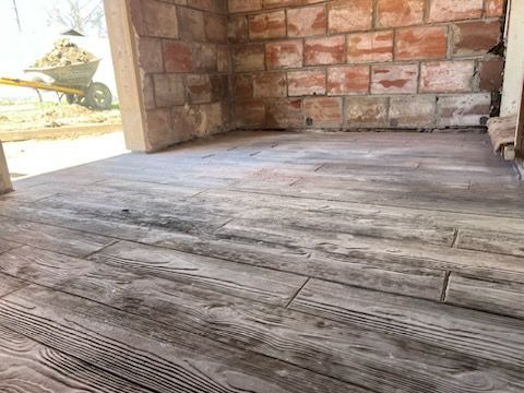 Weathered wooden porch floor beside a red brick wall, with sunlight and a glimpse of garden outside.