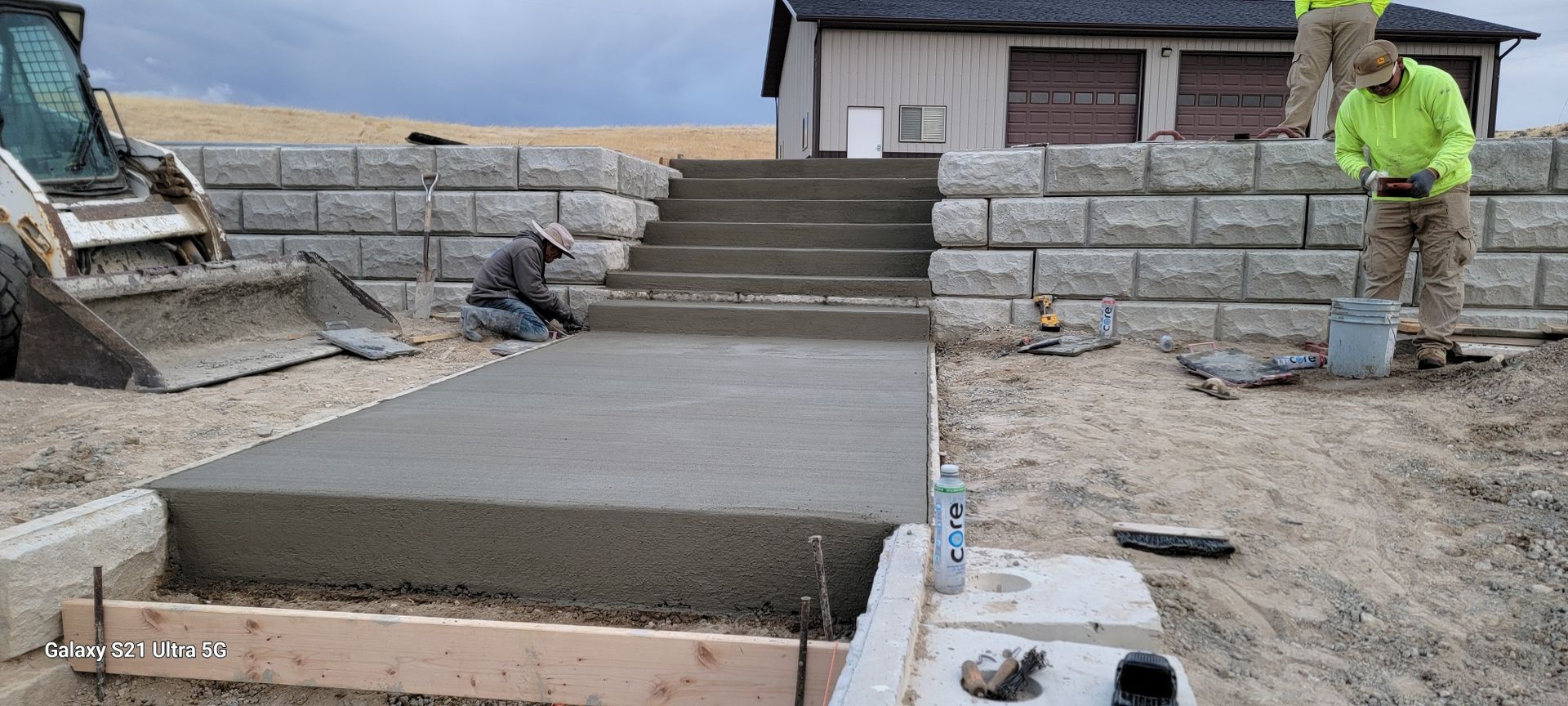 Concrete walkway and steps under construction beside stone retaining walls, with a worker in a neon jacket.