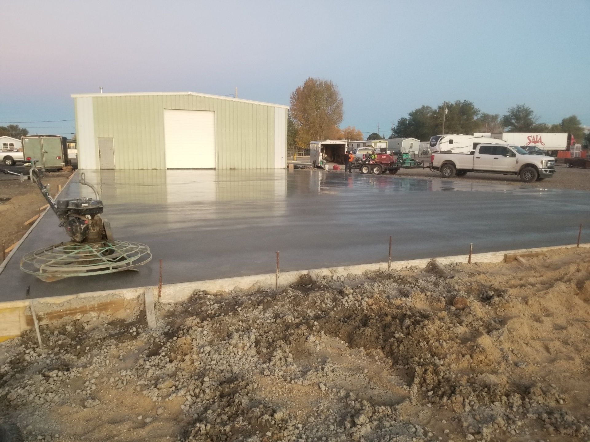 Concrete lot with a pale metal building, parked work trucks, and construction equipment in the foreground.