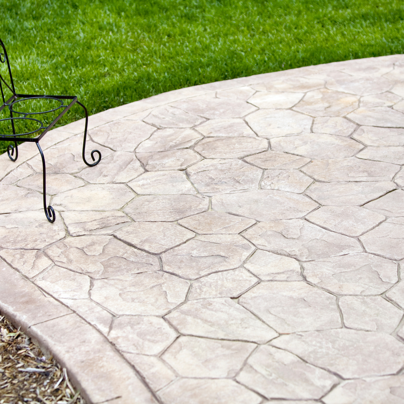 Curved stamped concrete patio beside green lawn, with part of a wrought-iron table visible.
