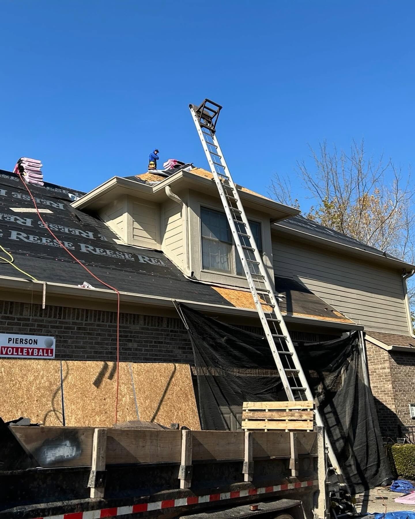 Roofing work on a two-story house with ladders and tarps under a clear blue sky