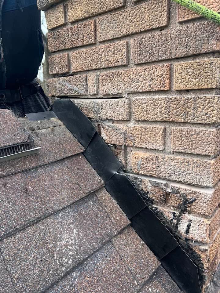 Damaged roof flashing along the seam between shingles and brick wall, with black sealant visible.