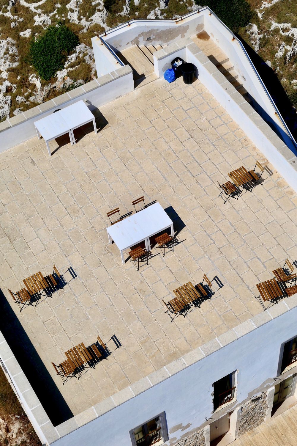 Rooftop terrace with white tables and chairs, viewed from above, beside a white building and rocky ground.